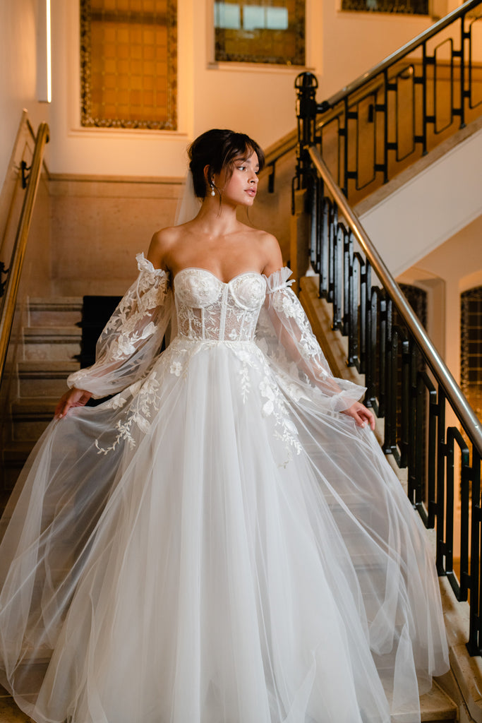 JOY is a real head-turner! This dreamy wedding gown is overflowing with romance and femininity. The luscious silhouette is brought to life with a daring low-back corset and a full cloche skirt. Pompous off-the-shoulder sleeves are crafted from contemporary lace, while a matching veil completes the look.