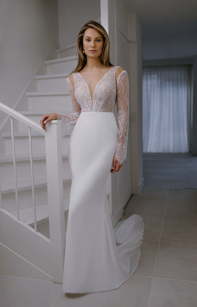 IYAR is a sexy and sophisticated Wedding Dress with a classic cut. The deep V-neckline is embellished with floating pearls. The gentle lace top features a peek-a-boo cold shoulder that reveals some extra skin. The long chiffon skirt boasts an elegant triangle train. The look is finished off with hand-sewn buttons that run down the low-cut back.