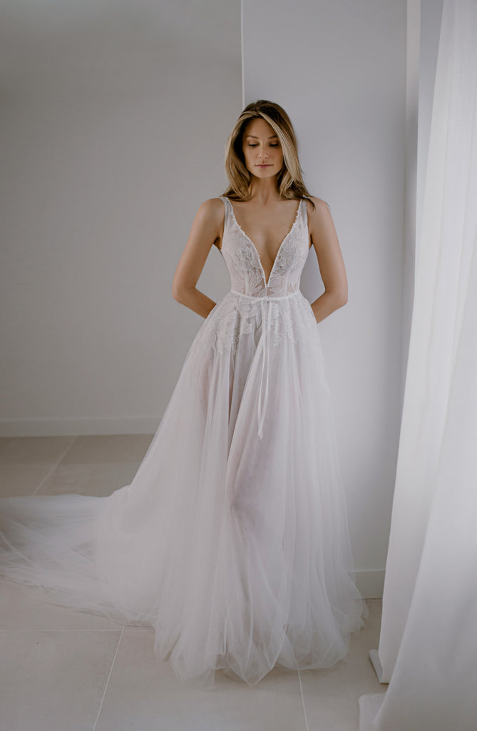 APRIL is a soft, ultra-romantic Wedding Dress that is as fresh as spring. APRIL is a full cloche tulle dress with a long train and a seductive plunge neckline that is adorned with dainty pearls. The top boasts a dreamy lace floral pattern that draws the eye to a petit bowed belt.  Complete the look with a long, dramatic veil, and a modern lace overtop, sold separately. The overtop features irresistible puffy sleeves that end with a manchette.