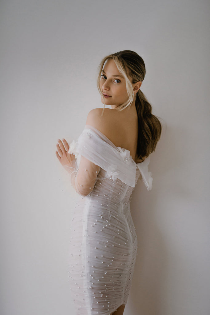 BERLINA is a short and sweet bridal dress that frames the body beautifully. BERLINA is crafted in gorgeous, draped tulle, that is embellished with delicate pearls throughout. The off-the-shoulder sleeves are adorned with 3D lace flowers, for a soft accent on this form-fitting dress.