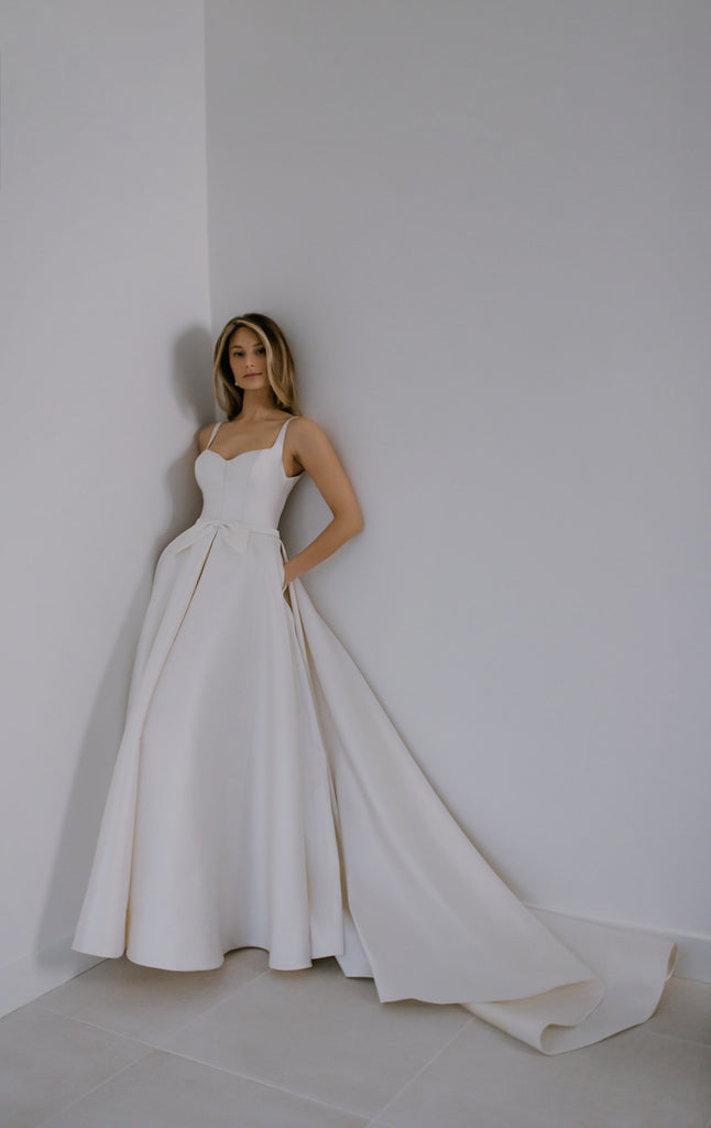PEARLA takes the classic fairytale princess dress and gives it a contemporary twist. Crafted from top-of-the-line Mikado Silk, this gown is structured and smooth, which makes it appear as if it’s floating around the bride. PEARLA features a romantic sweetheart neckline, a sexy low back, deep folds throughout the skirt, and a naïve bow to embellish the delicate belt.