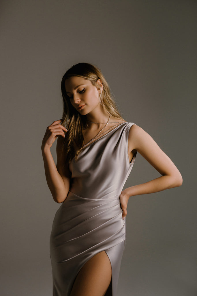 Roll out the red carpet for RONA! This asymmetric dress features a high slit and beautiful draping that flows with the body. The grey-silver satin gives this dress a statuesque, almost Grecian quality. The flattering fit is finished off with a crystal trim, ensuring you’ll feel like a star of the silver screen.