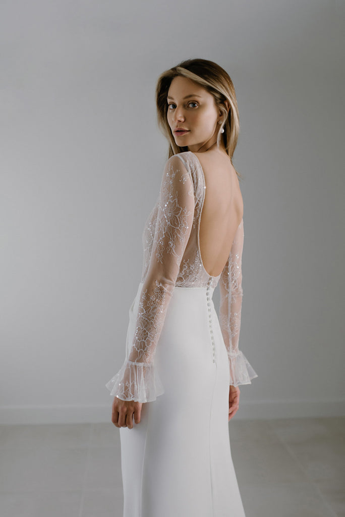 YULI is a modern beauty, with a crisp and clean appearance. The top of this bridal dress is crafted in beautiful 3D lace that sparkles lightly, and the sleeves are tapered with a romantic Volan. The draped folded skirt features a seductive side slit, and button details that run down the back. YULI can be layered with a matching sparkly 3D lace overskirt that gives this bridal gown a dramatic and alluring train.
