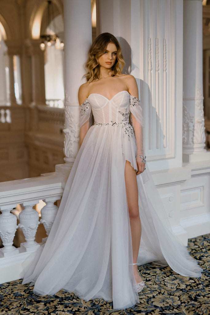 BLANCHE is a timeless Wedding Dress. It features a handmade drape corset, romantic sleeves, and a gathered layered tulle skirt with a high slit and intriguing volan, making it a true eye-catcher. This dress is adorned with hand-embroidered lace, speckled with light-catching sequins and dark blue stones.  BLANCHE is the ultimate gown for a bride that walks the line between fairy-tale and fierce.
